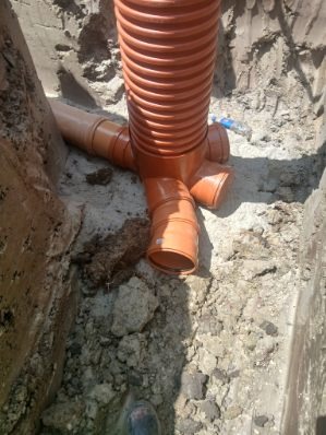 Pomarańczowe rury kanalizacyjne z trójnikiem w wykopie, widoczne połączenia. Instalacja w trakcie montażu, ziemia wokół rur.