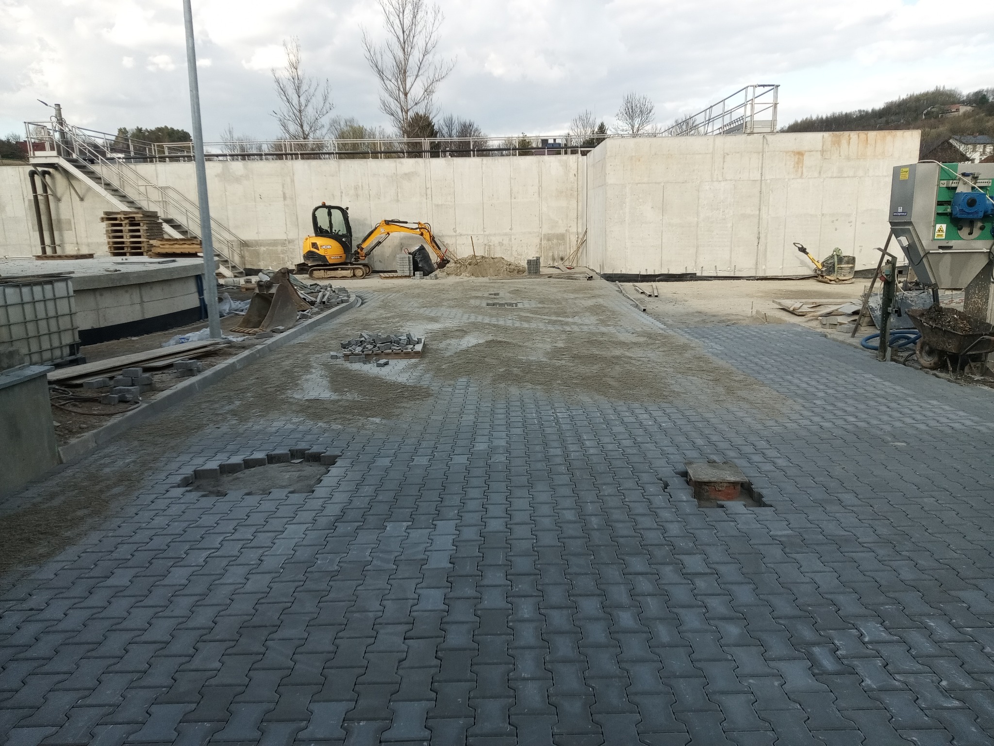 Częściowo ułożona, szara kostka brukowa na placu budowy. W tle widoczny mały, żółty koparka, betonowy mur i maszyny budowlane. Dwa puste miejsca po kostce.