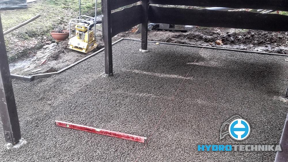 Przygotowanie podłoża pod taras: ubity tłuczeń, poziomica, zagęszczarka w tle, widoczne obrzeża i metalowe słupy konstrukcji tarasu. Logo firmy w prawym dolnym rogu.