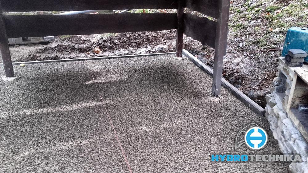 Przygotowany teren pod budowę altany, wysypany drobnym szarym kruszywem, obrzeża wyznaczone krawężnikami, widoczny fragment murowanej konstrukcji i logo firmy.