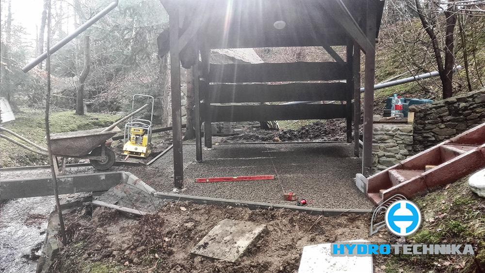 Przygotowanie podłoża pod konstrukcję w ogrodzie: ubity żwir, w tle taczka, zagęszczarka, poziomica i narzędzia, widoczne też schody i logo firmy.