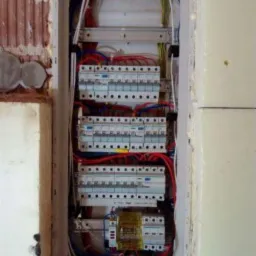 Otwarta skrzynka bezpiecznikowa z widocznymi bezpiecznikami, przewodami w kolorach niebieskim, czerwonym, białym i żółto-zielonym, zamontowana w ścianie w trakcie remontu.