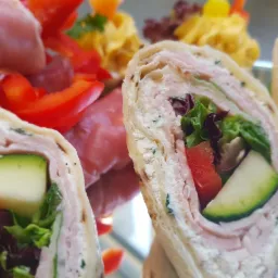 Tortilla wrap z szynką, serem, ogórkiem, papryką i sałatą na lustrzanej tacy, z widocznymi w tle przekąskami.