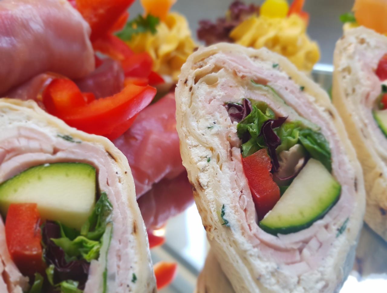 Tortilla wrap z szynką, serem, ogórkiem, papryką i sałatą na lustrzanej tacy, z widocznymi w tle przekąskami.