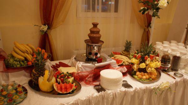 Stół z fontanną czekoladową, owocami (banany, winogrona, ananasy, arbuzy, truskawki) i babeczkami, udekorowany białym obrusem i materiałami w odcieniach czerwieni i złota.