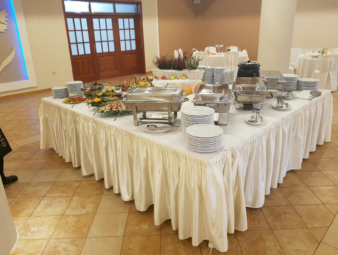 Stół bufetowy z potrawami, naczyniami i dekoracjami na przyjęciu, widoczne podgrzewacze i talerze, w tle stoliki z białymi obrusami.