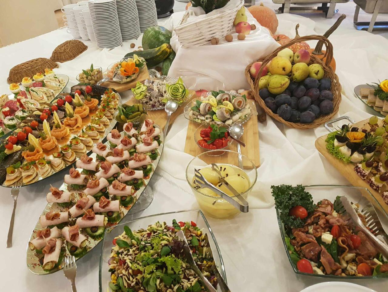 Bogato zastawiony stół cateringowy z różnorodnymi przekąskami, sałatkami, owocami i warzywami, w tym roladki z szynki, koreczki, sałatki w szklanych misach i kosz ze śliwkami i gruszkami, całość...