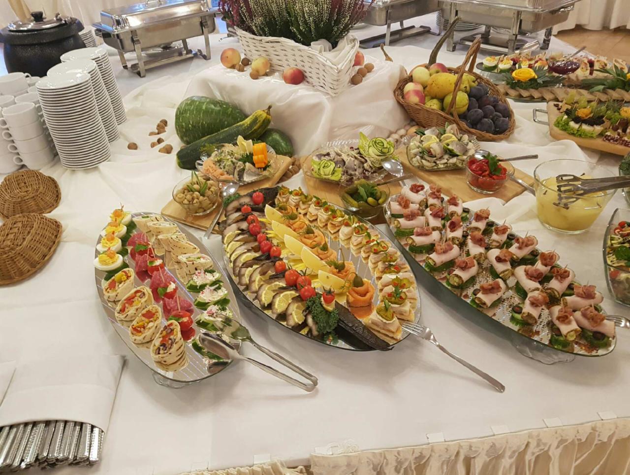 Bogato zastawiony stół z przekąskami: koreczki, sałatki, owoce, warzywa, oraz podgrzewacze do potraw, całość na białym obrusie.