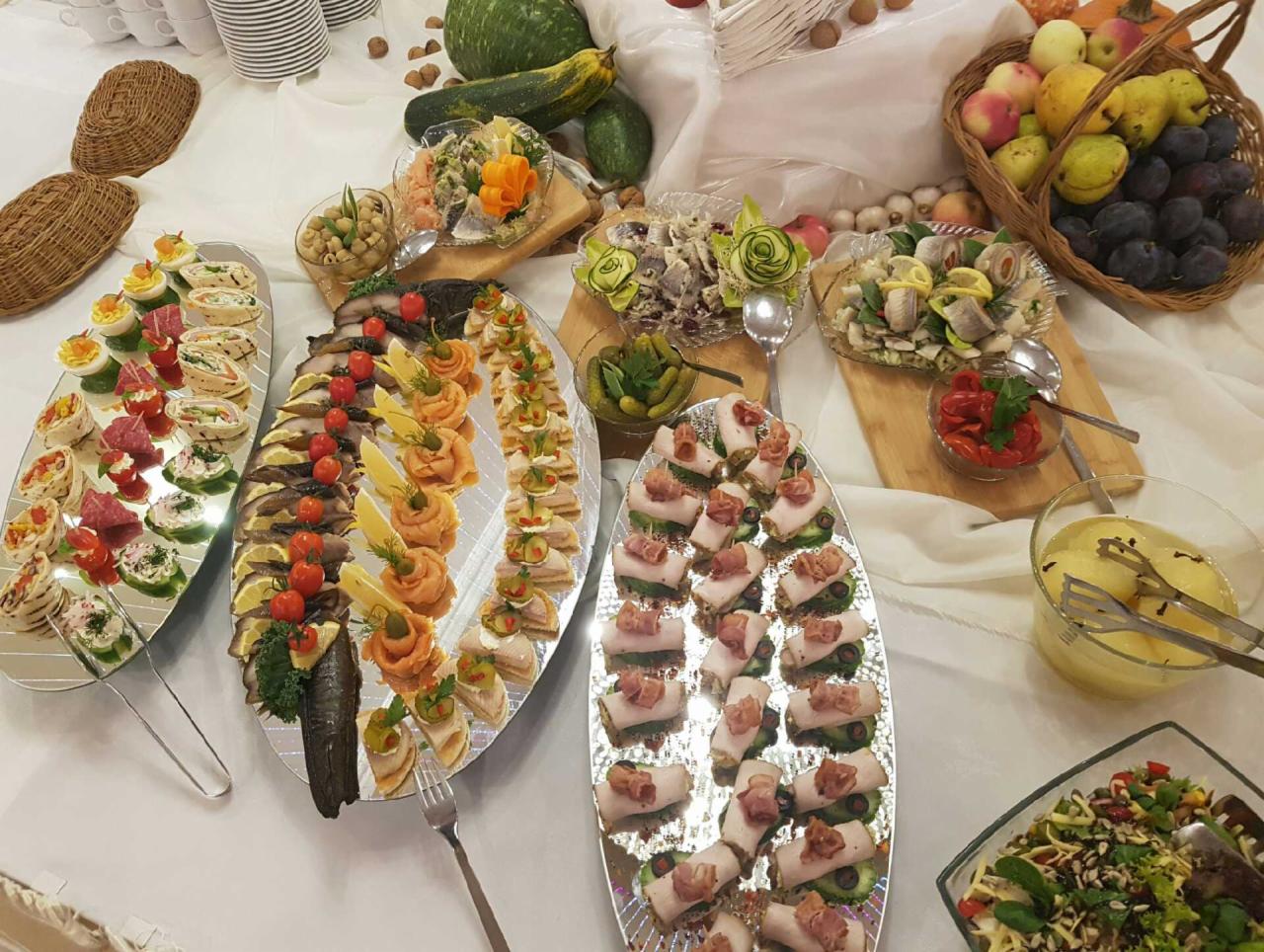 Bogato zastawiony stół z różnorodnymi przekąskami na srebrnych paterach, w tle kosz z owocami i dekoracyjne warzywa.