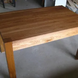 Stół z drewna o jasnym blacie z widocznym usłojeniem i ciemniejszych nogach, na szarym betonowym podłożu, w tle materiały budowlane.