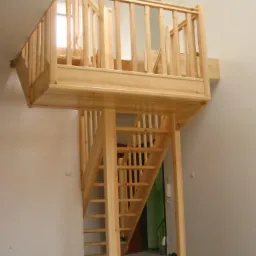 Drewniane schody prowadzące na antresolę z balustradą, wykonane z jasnego drewna w surowym stanie, widok z dołu na konstrukcję w jasnym pomieszczeniu z zielonymi drzwiami w tle.