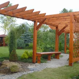 Drewniana konstrukcja z ławką w ogrodzie, widoczna pergola z kratownicą, w tle altana i zieleń. Ujęcie z poziomu gruntu, widoczny fragment ścieżki z kamienia.
