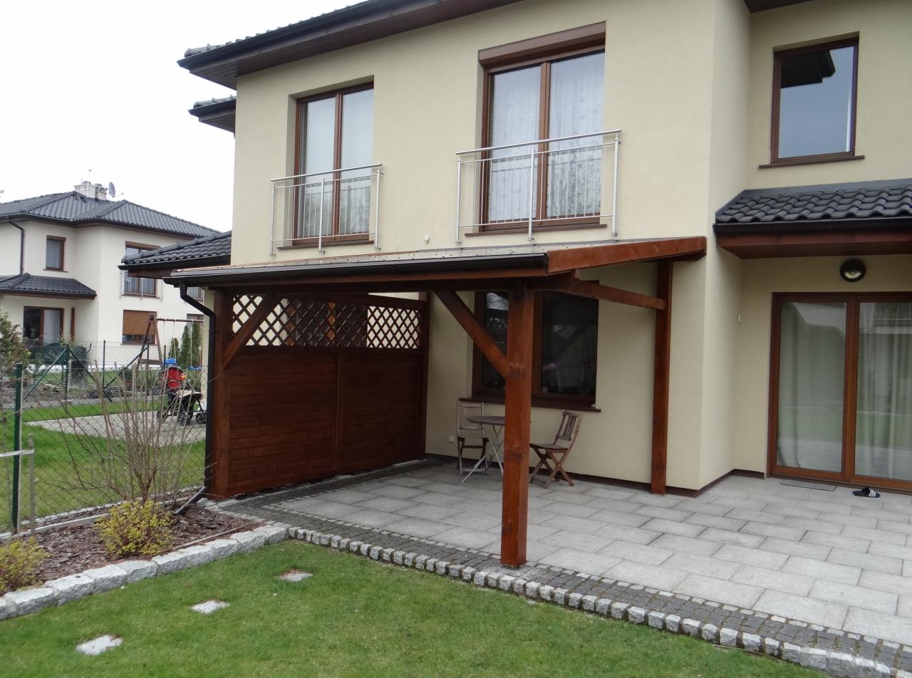 Drewniana konstrukcja z ażurową ścianką przylegająca do domu, tworząca zadaszone miejsce wypoczynku z kamienną podłogą i dwoma krzesłami.