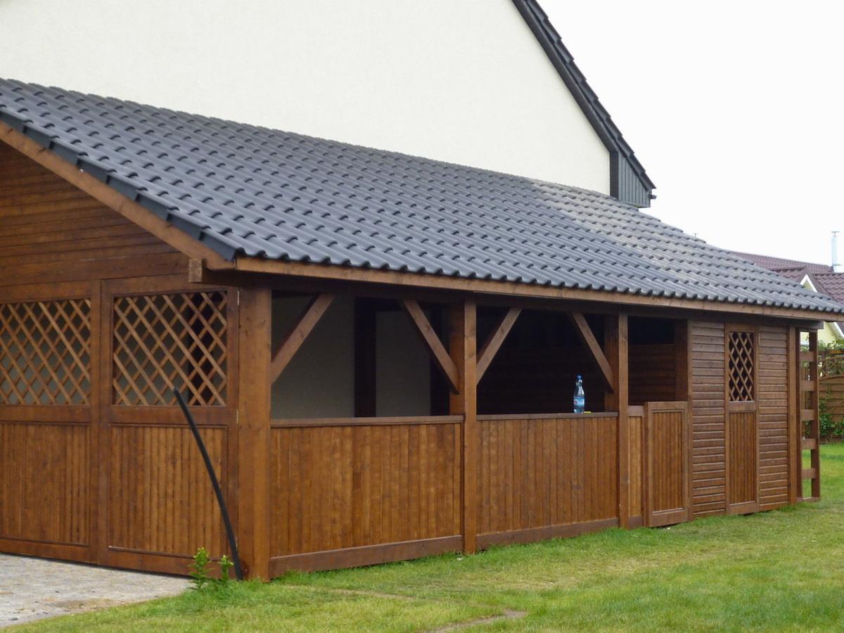 Drewniana altana z ciemnym, skośnym dachem krytym dachówką, z kratownicowymi panelami bocznymi i butelką wody na parapecie.