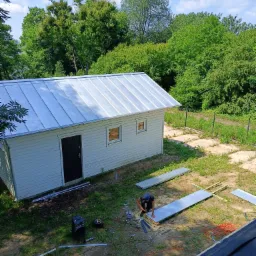 Prace dekarskie przy małym białym budynku z blachy trapezowej na dachu, widok z góry, dekarz w trakcie montażu elementów, rozłożone narzędzia i materiały budowlane na trawie.