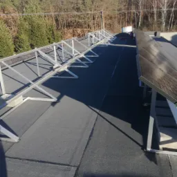 Przygotowanie do montażu paneli słonecznych na płaskim dachu pokrytym papą, widoczne metalowe konstrukcje wsporcze i częściowo zamontowane panele.