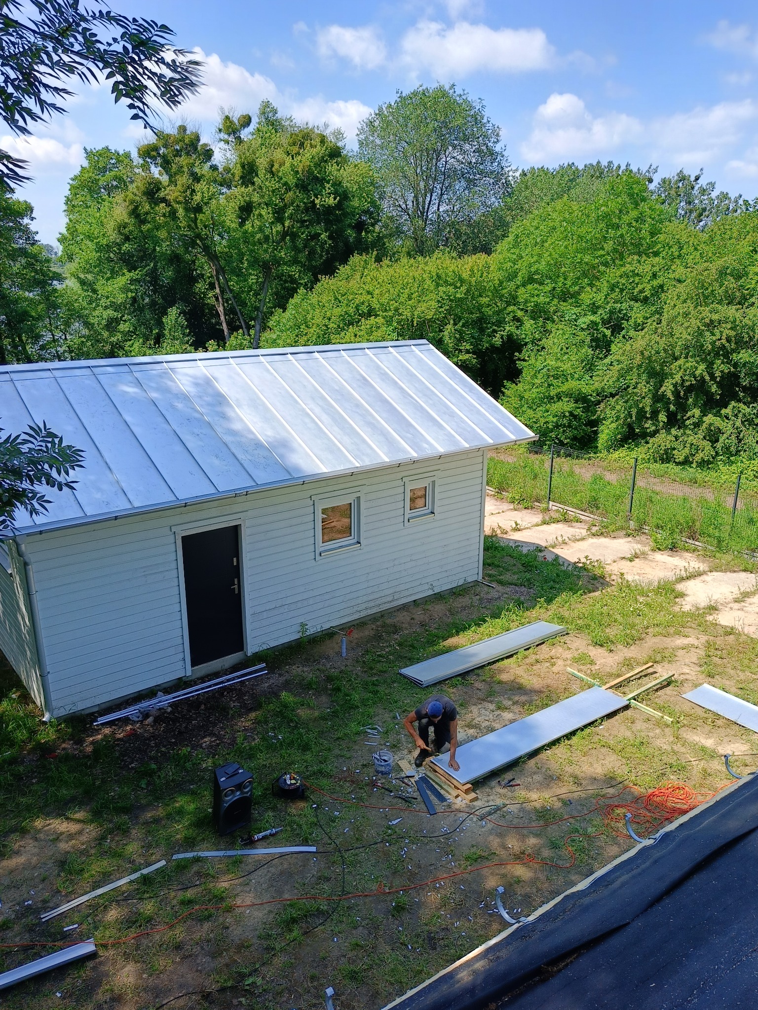 Prace dekarskie przy małym białym budynku z blachy trapezowej na dachu, widok z góry, dekarz w trakcie montażu elementów, rozłożone narzędzia i materiały budowlane na trawie.