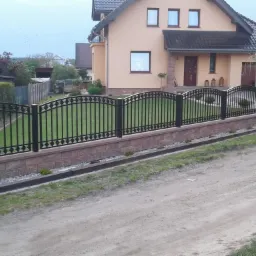 Czarne, metalowe ogrodzenie z dekoracyjnymi elementami, osadzone na murku z cegły klinkierowej, na tle domu jednorodzinnego i kościoła w oddali.