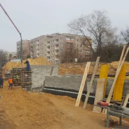 Budowa fundamentów budynku wielorodzinnego: pracownicy w odblaskowych kamizelkach, betonowy fundament z deskowaniem, pompa do betonu w tle, teren budowy z hałdami piasku.