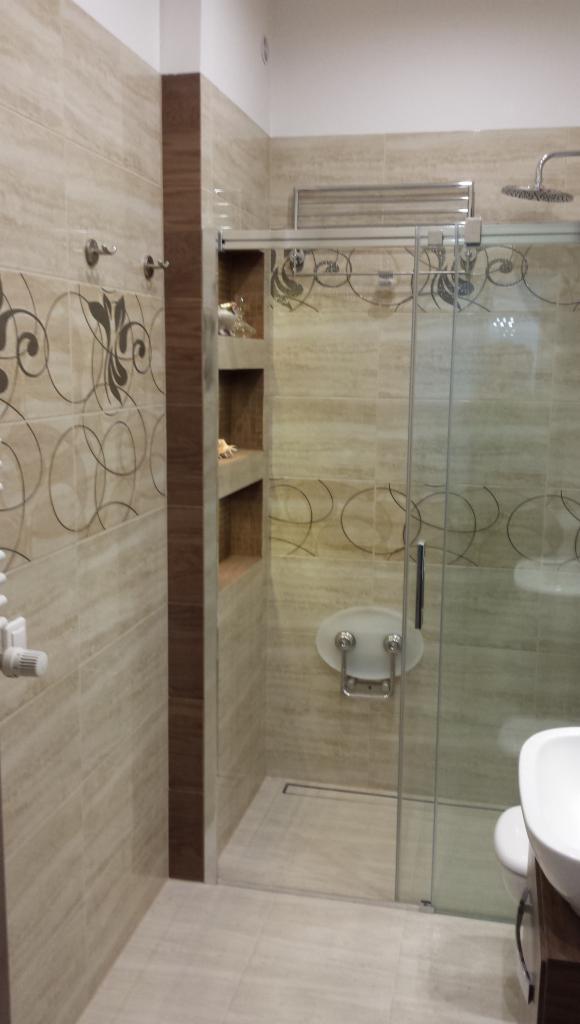 Wykończona łazienka z kabiną prysznicową ze szklanymi drzwiami przesuwnymi, beżowe płytki z motywem roślinnym, wbudowane półki z dekoracjami i odpływ liniowy w podłodze.