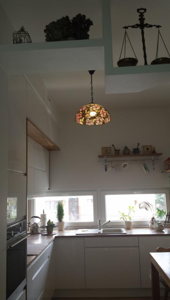Jasna kuchnia z białymi szafkami, drewnianym blatem i witrażową lampą wiszącą w stylu Tiffany, na półkach dekoracje.