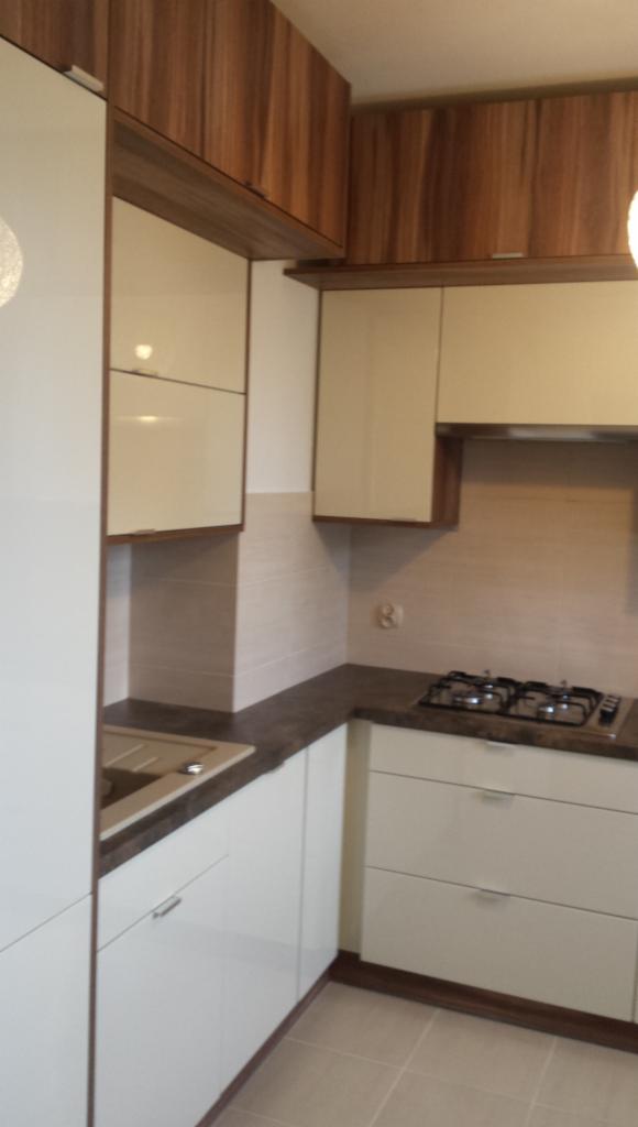 Nowoczesna kuchnia z białymi lakierowanymi szafkami dolnymi i górnymi, oraz drewnopodobnymi szafkami wiszącymi, beżowe płytki na ścianie i ciemny blat.