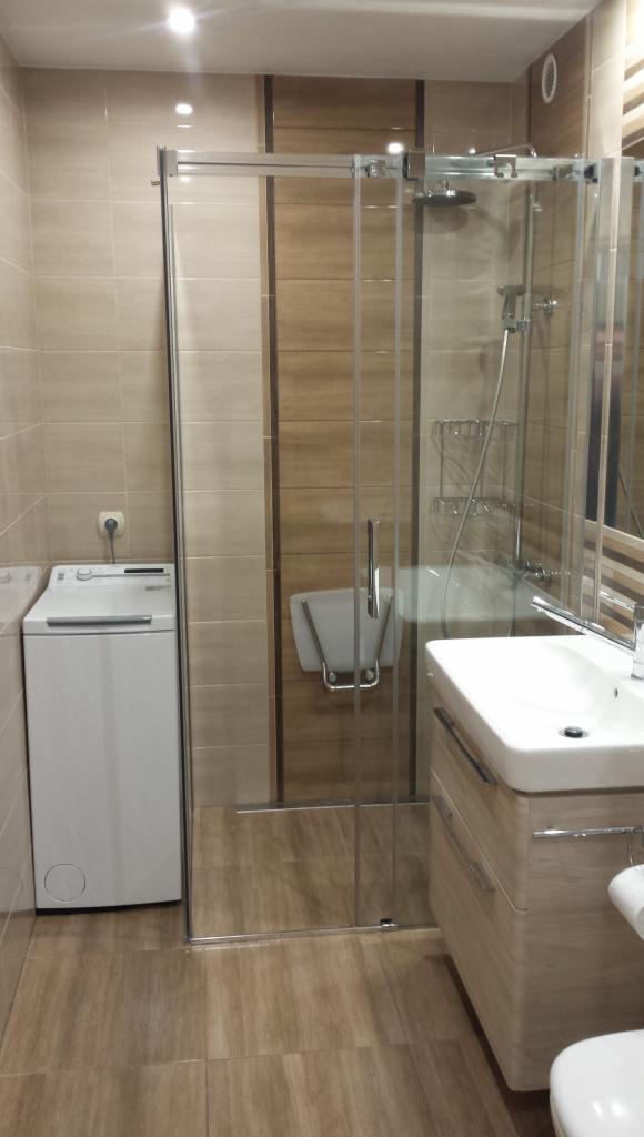 Nowoczesna łazienka z kabiną prysznicową typu walk-in, umywalką z szafką w odcieniach beżu i pralką, wykończona płytkami imitującymi drewno.