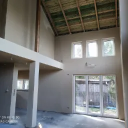 Wnętrze domu w trakcie wykańczania, świeżo położone jasne tynki na ścianach, widoczne drewniane elementy konstrukcyjne stropu i poddasza, duże okna i drzwi balkonowe wpuszczające naturalne światło.