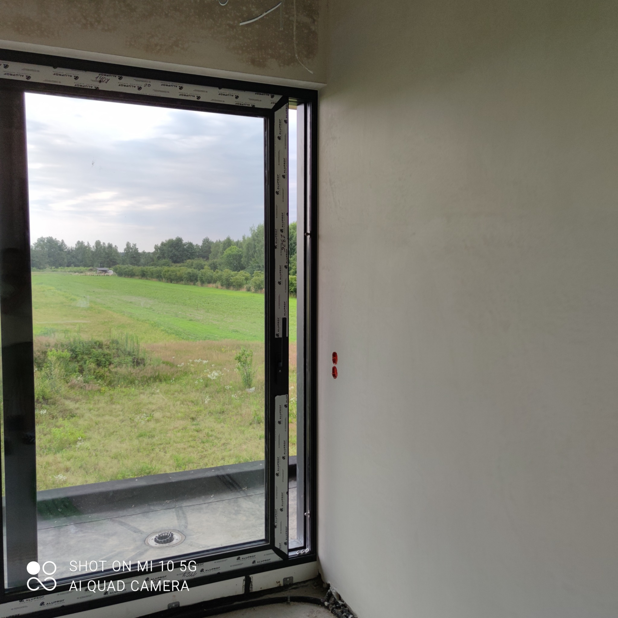 Świeżo otynkowana ściana wewnętrzna z widocznymi puszkami elektrycznymi, z oknem z czarną ramą Aluplast z widokiem na zielone pole i las w oddali.