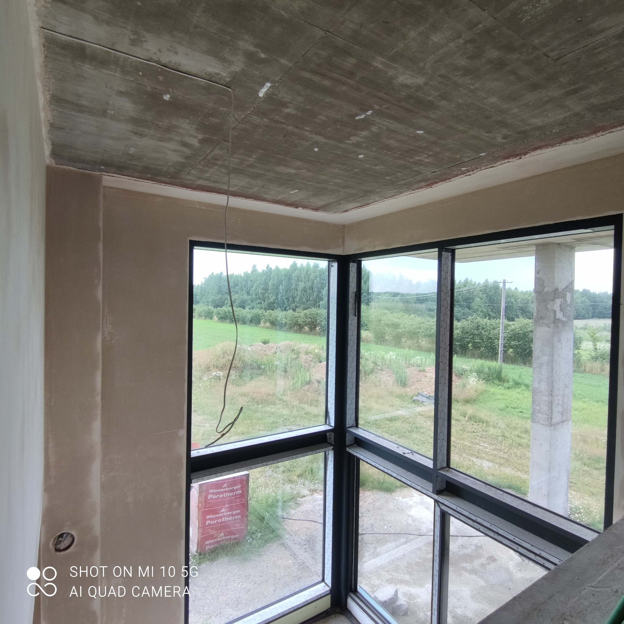 Surowy betonowy sufit z widocznymi śladami po szalunku i świeżo nałożone tynki na ścianach w narożnym pokoju z dużymi oknami wychodzącymi na zielony krajobraz.
