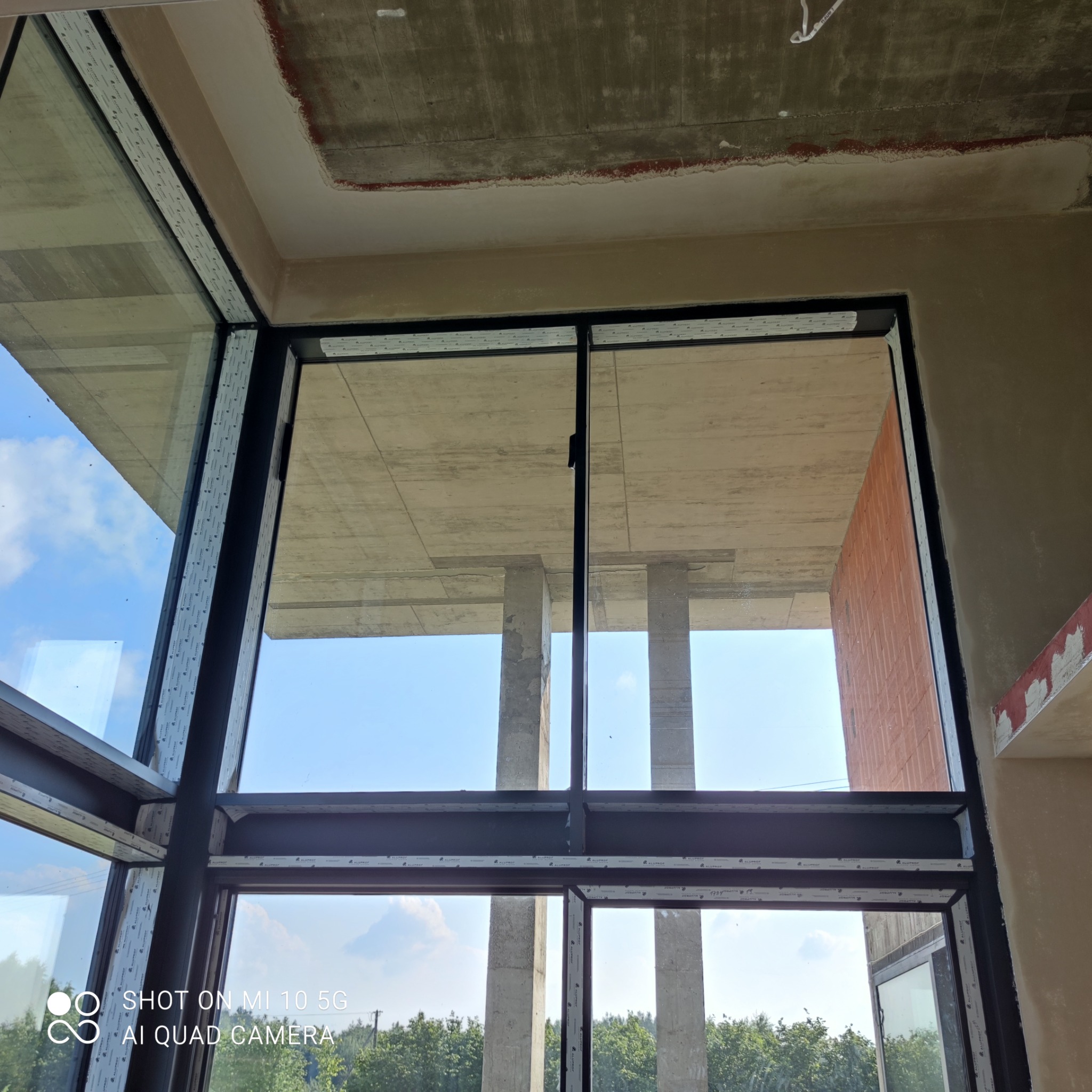 Wnętrze budynku w trakcie prac budowlanych, widoczne surowe ściany, betonowy strop i zamontowane okna z czarnymi ramami, przez które widać konstrukcję budynku i niebo.