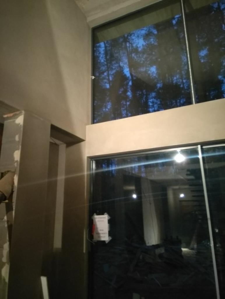 Surowe ściany po nałożeniu tynku w pomieszczeniu z dużymi oknami, widoczne odblaski drzew na szybach.