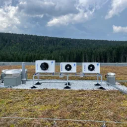Trzy jednostki zewnętrzne klimatyzacji na dachu pokrytym roślinnością i białym żwirem, widok na las i pochmurne niebo.