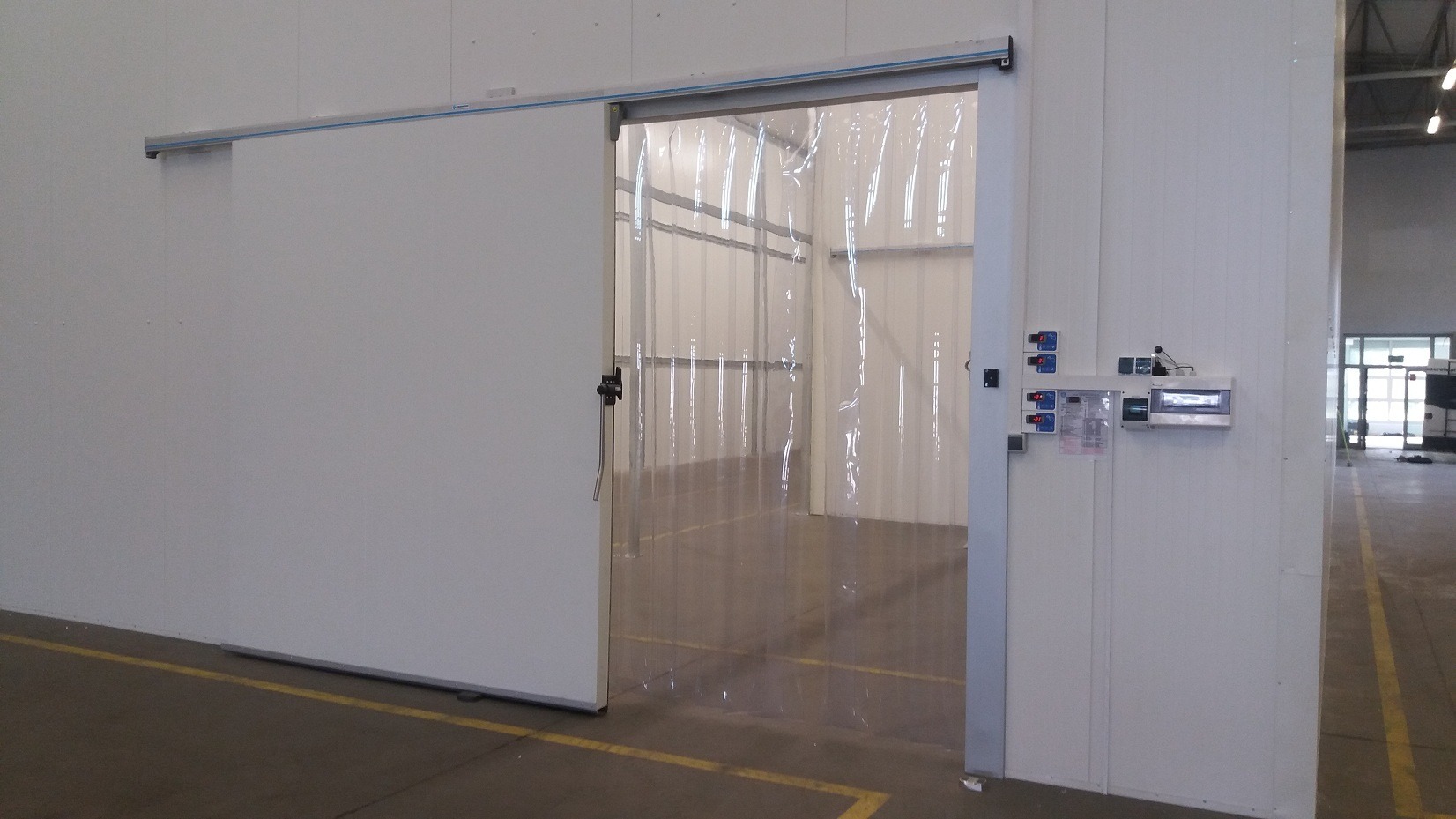 Przesuwne drzwi do chłodni z kurtyną paskową z PCV, widoczne regulatory temperatury na ścianie, podłoga z żółtymi liniami.