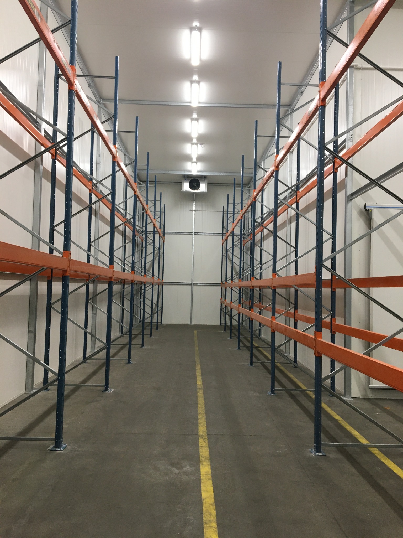 Puste metalowe regały magazynowe z pomarańczowymi poprzeczkami w chłodni, widoczna linia wyznaczająca strefę na posadzce i wentylator na suficie.