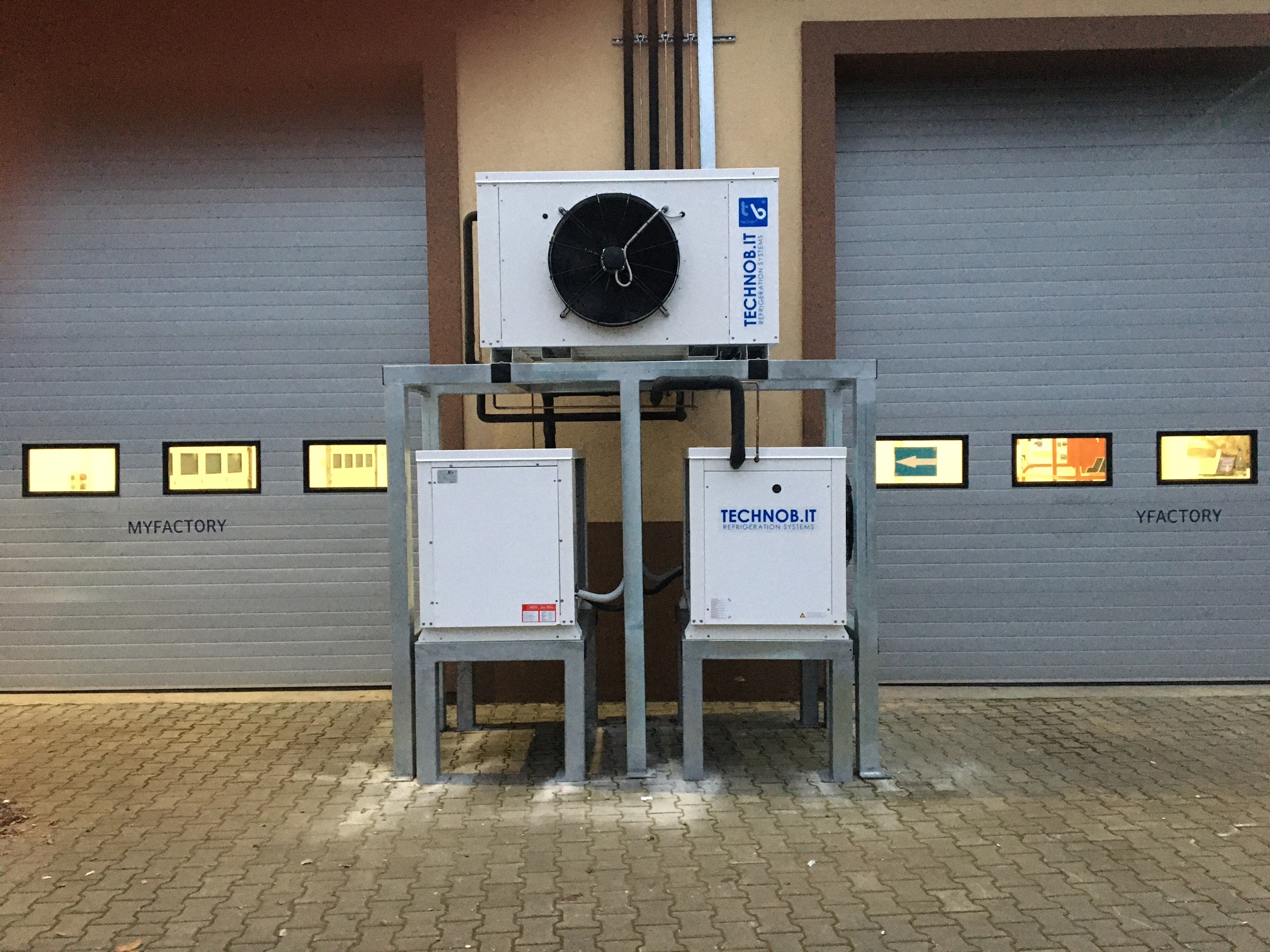 Instalacja trzech jednostek chłodniczych Technob.it na metalowej konstrukcji przed szarą bramą garażową z oknami w fabryce MYFACTORY i YFACTORY