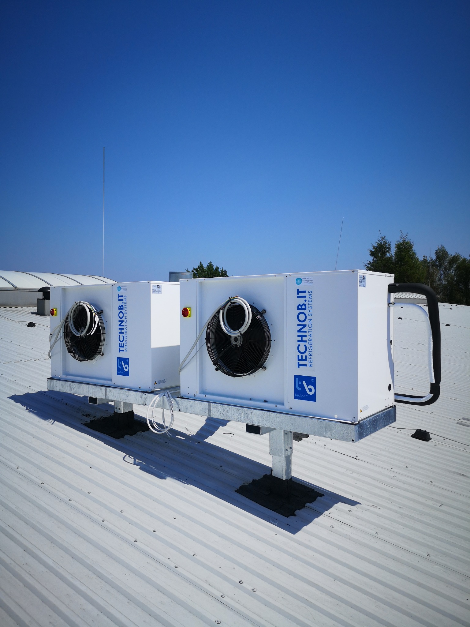 Dwa agregaty chłodnicze marki TECHNOb.IT zamontowane na dachu z blachy trapezowej, widoczne przewody i elementy montażowe.