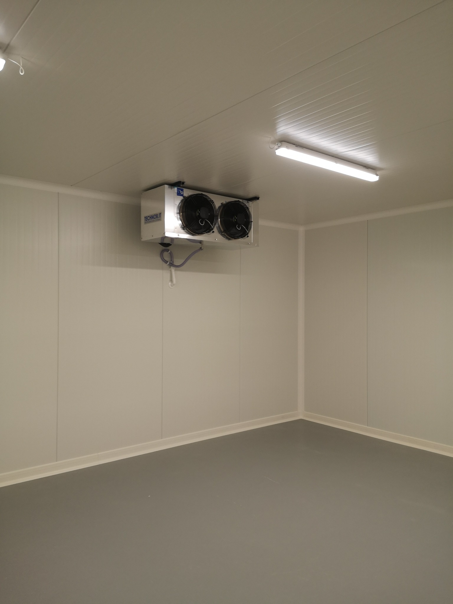 Wnętrze chłodni z szarym podłożem, białe ściany z paneli, oświetlenie jarzeniowe i zamontowany pod sufitem agregat chłodniczy marki TECHNObit z dwoma wentylatorami.