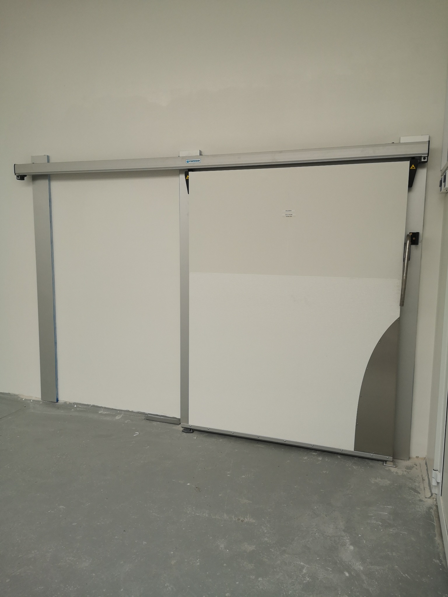 Przesuwne drzwi chłodnicze w komorze chłodniczej, widoczne szyny i uchwyt, z dolną krawędzią drzwi wykończoną metalową osłoną w kształcie łuku.