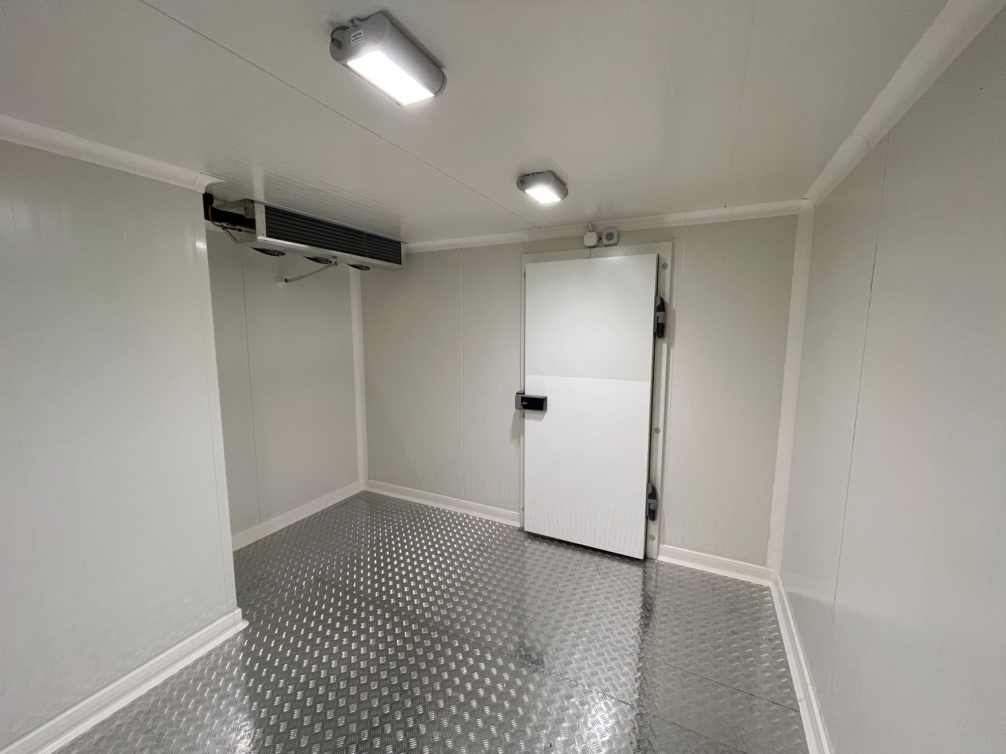 Wnętrze chłodni z metalową podłogą, widoczny agregat chłodniczy pod sufitem i białe panele ścienne.