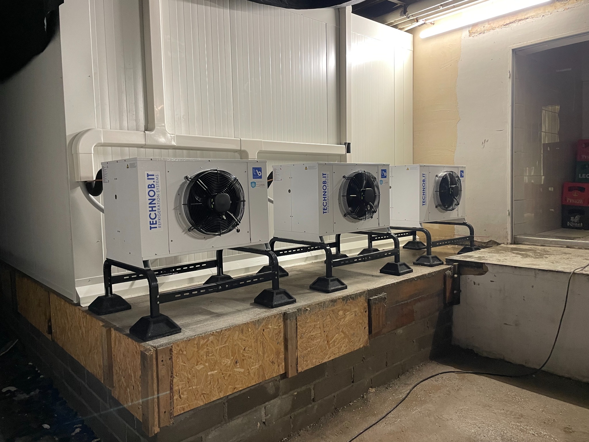 Trzy agregaty chłodnicze marki TECHNOb.IT zamontowane na podwyższeniu z płyt OSB w pomieszczeniu magazynowym, widoczne rury instalacyjne i kartony piwa w tle.