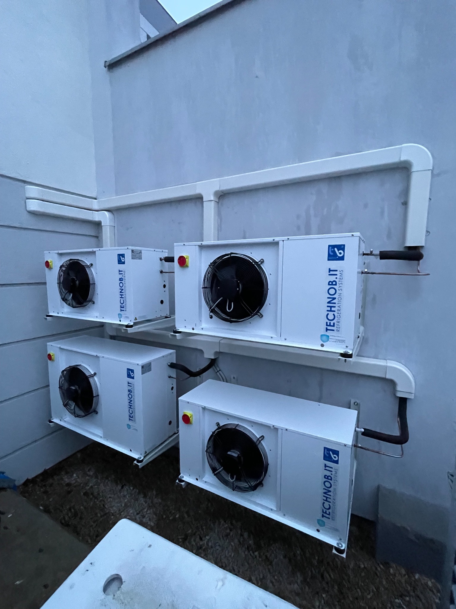 Cztery białe jednostki chłodnicze marki TECHNOb.IT zamontowane na zewnątrz budynku, z widocznymi przewodami i wentylatorami, na tle szarej ściany i żwirowego podłoża.