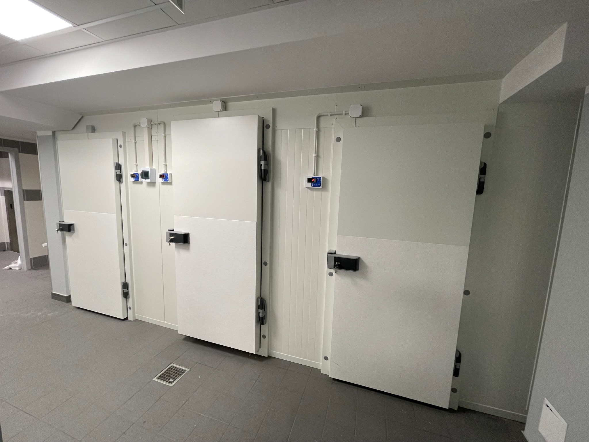 Wnętrze chłodni z trzema białymi drzwiami, widoczne regulatory temperatury i kratka odpływowa w podłodze.