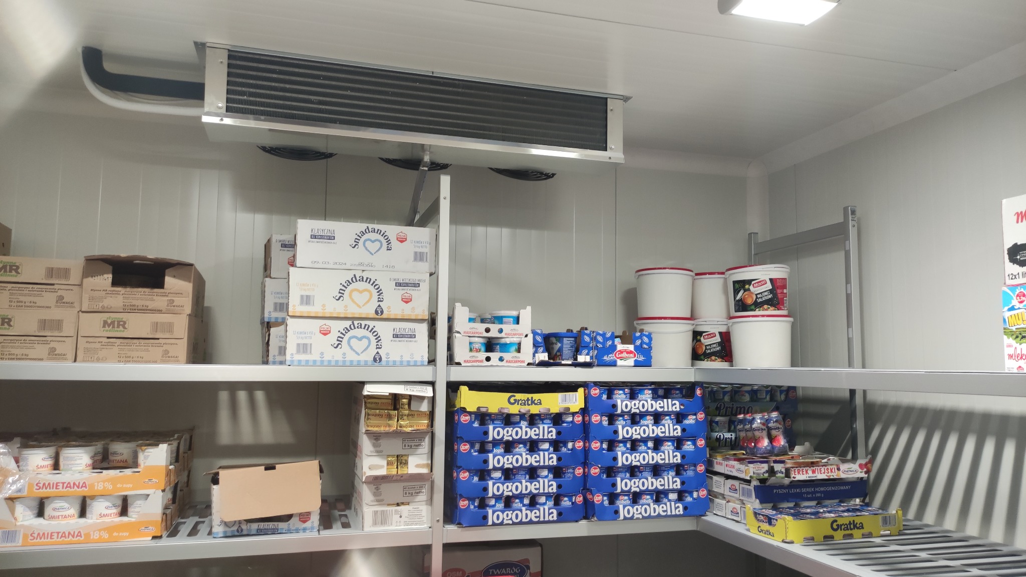 Wnętrze chłodni z półkami wypełnionymi produktami mlecznymi: śmietana, jogurty, twarogi, serki homogenizowane, widoczny parownik chłodniczy pod sufitem.