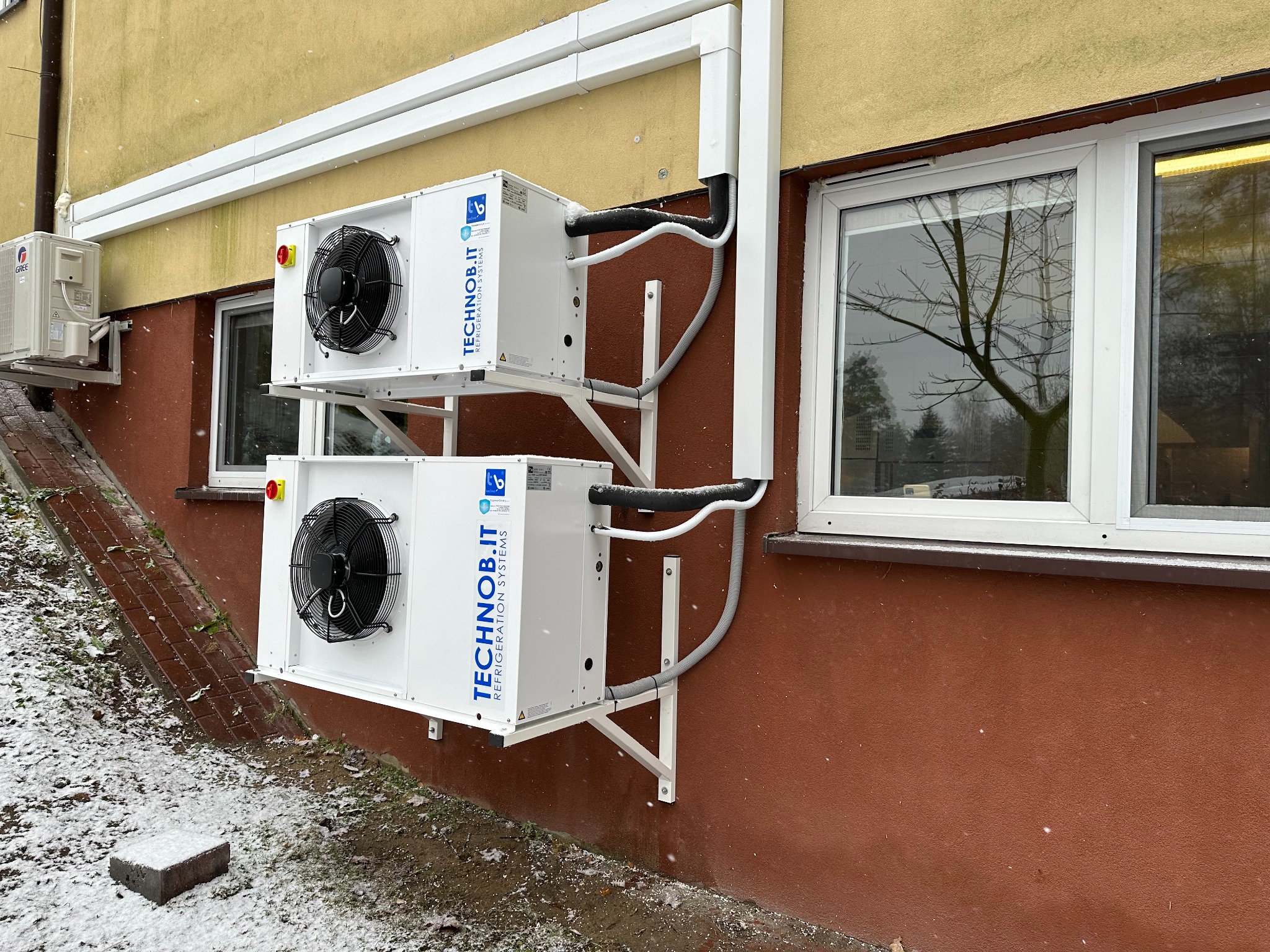 Dwa agregaty chłodnicze marki TECHNOb.IT zamontowane na wspornikach na zewnątrz budynku, z widocznymi przewodami i elementami instalacji odprowadzającymi skropliny, w zimowej scenerii z padającym...