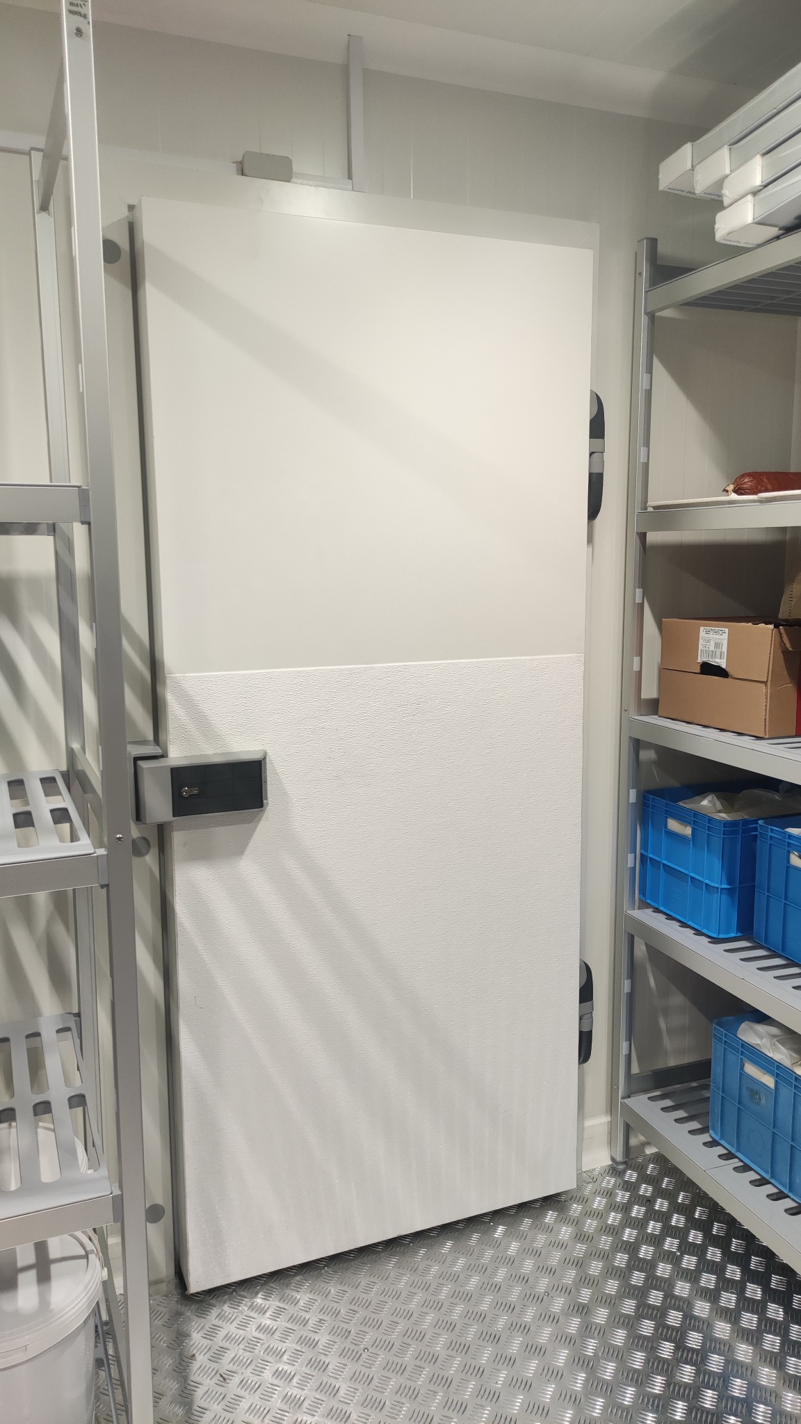 Wnętrze chłodni z białą, dwuczęściową bramą z zamkiem, metalowe regały z niebieskimi pojemnikami i kartonowym pudełkiem, podłoga z blachy ryflowanej.