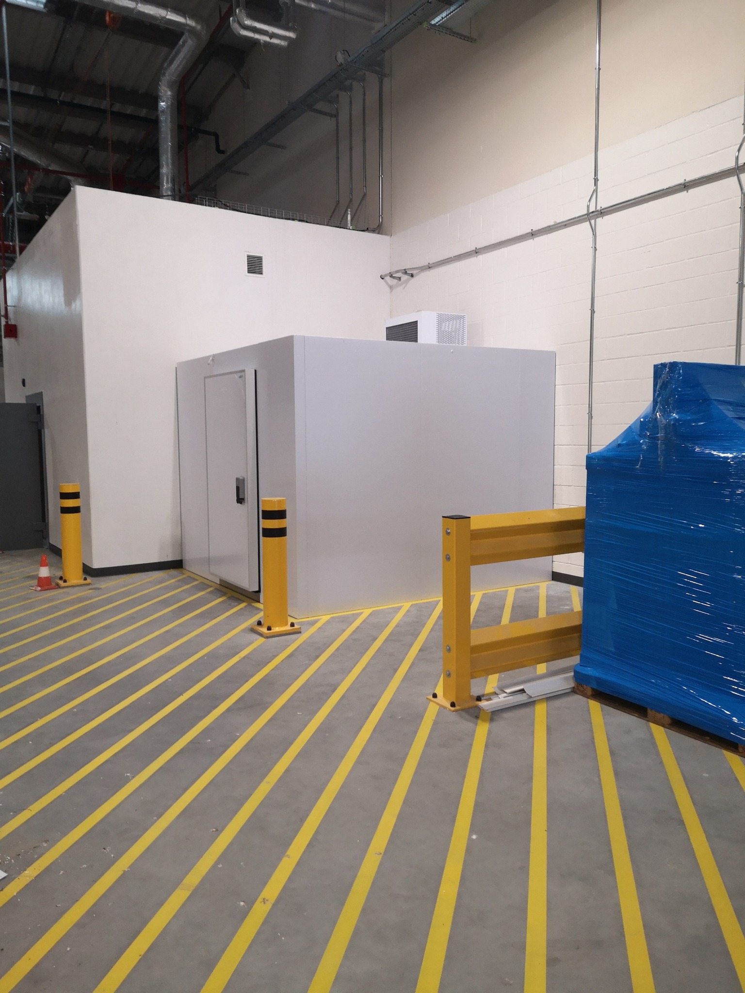 Nowoczesna komora chłodnicza z drzwiami, zabezpieczona żółtymi słupkami i barierkami, w tle instalacje przemysłowe pod sufitem.