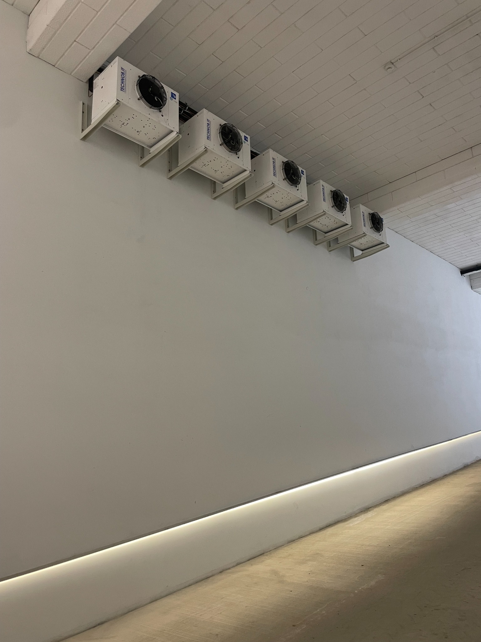 Rząd sześciu białych agregatów chłodniczych marki Technob.it zamontowanych pod sufitem w chłodnym, minimalistycznym wnętrzu z betonową podłogą i subtelnym oświetleniem przypodłogowym.