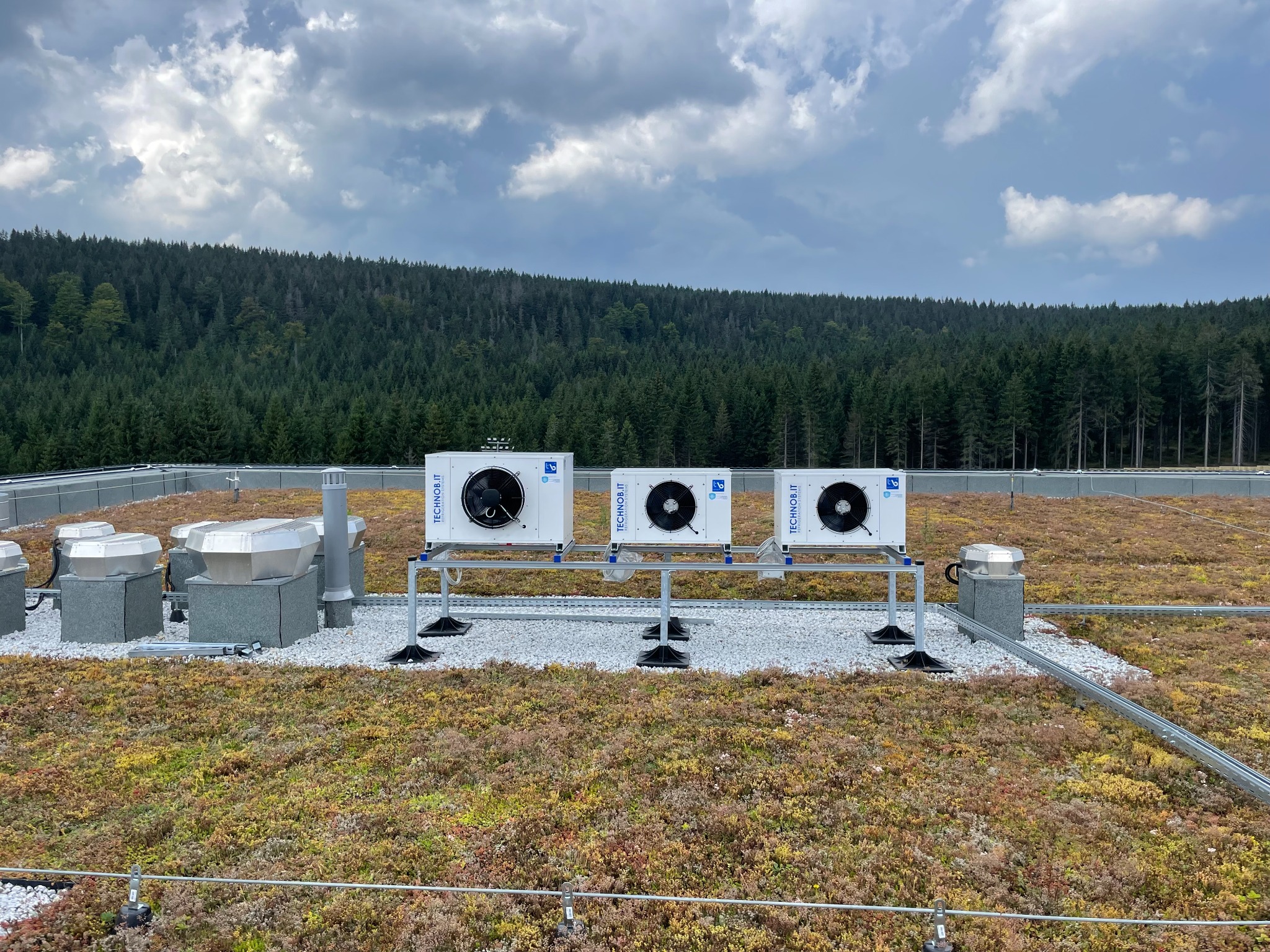Trzy jednostki zewnętrzne klimatyzacji na dachu pokrytym roślinnością i białym żwirem, widok na las i pochmurne niebo.