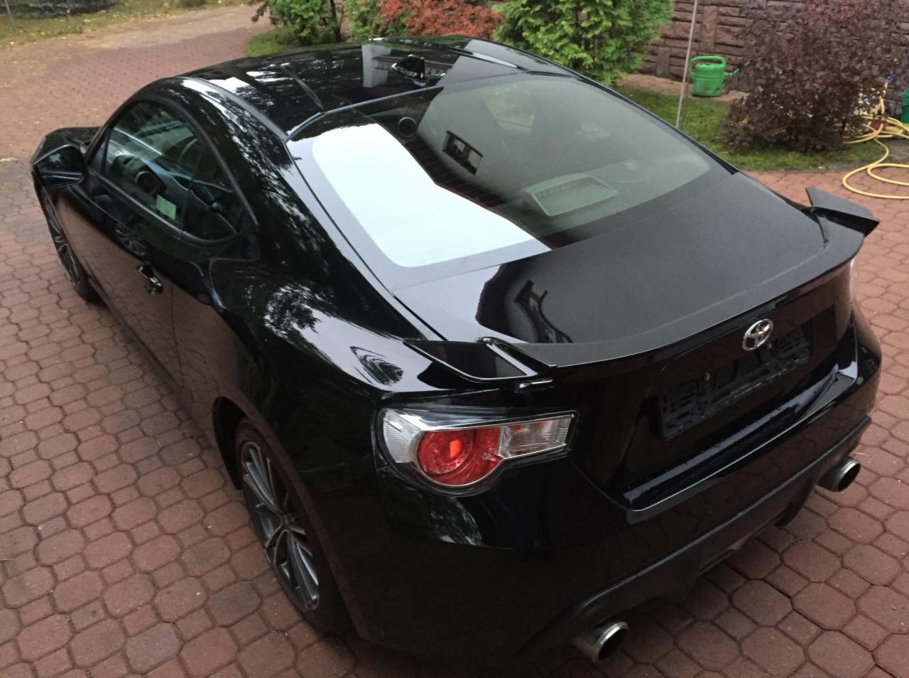 Błyszczący, czarny samochód sportowy Toyota GT86 z widocznym spojlerem na tylnej klapie, zaparkowany na czerwonej kostce brukowej, z zielenią w tle.